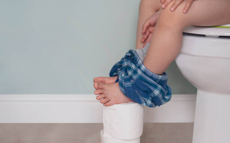 child sitting on toilet with feet on toilet paper rolls