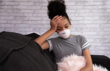 Black adolescent with hand pressed to forehead and wearing a mask