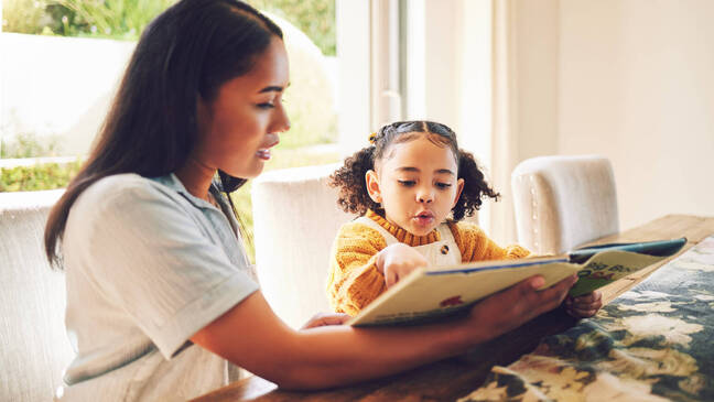 Woman and young girl sitting at table looking at book.