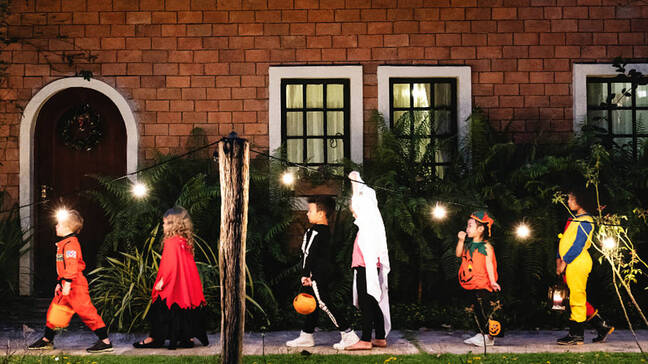 Kids walking in a line, dressed up in costumes. It is nighttime and they are walking in front of a house.